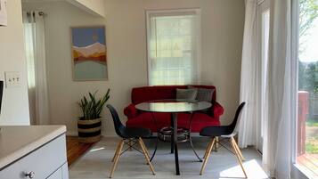 Una habitación con un sofá rojo, una mesa redonda y dos sillas. Hay un ventilador de techo, una ventana con persianas y un cuadro en la pared.