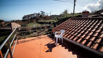 A terrace with a white plastic chair, a tiled roof, and a view of a hilly landscape.