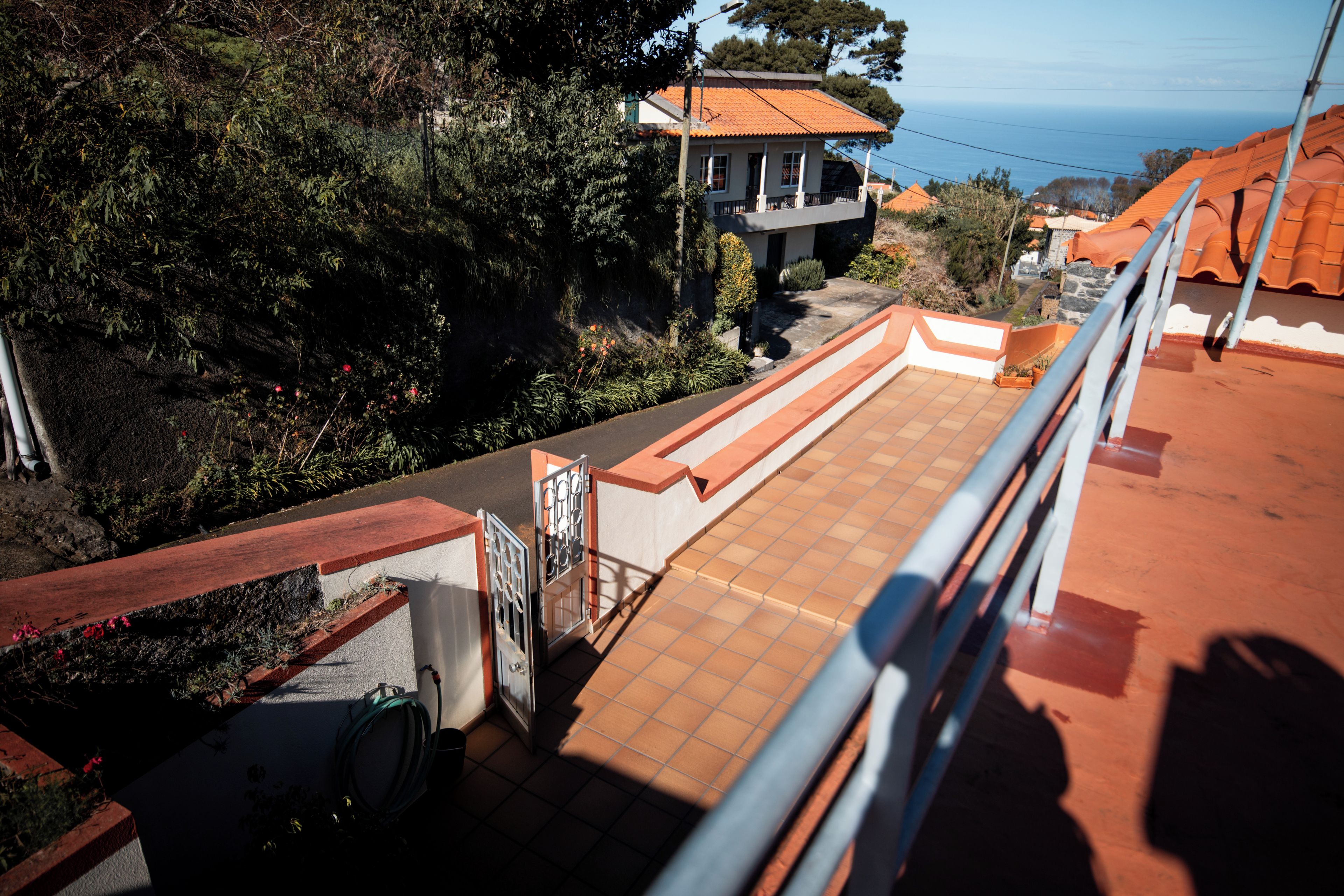 A rooftop terrace with a view of the sea, a garden, and a building with a tiled roof.