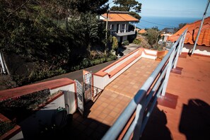 A rooftop terrace with a view of the sea, a garden, and a building with a tiled roof.