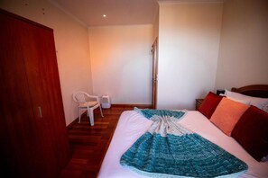 Un dormitorio con cama, un armario de madera, una silla blanca y una mesita de noche con lámpara.