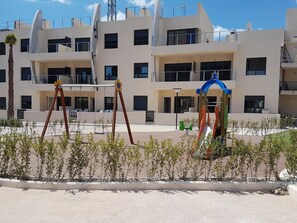 Um edifício moderno com várias varandas, um parquinho com balanços e escorregadores, e um céu claro.
