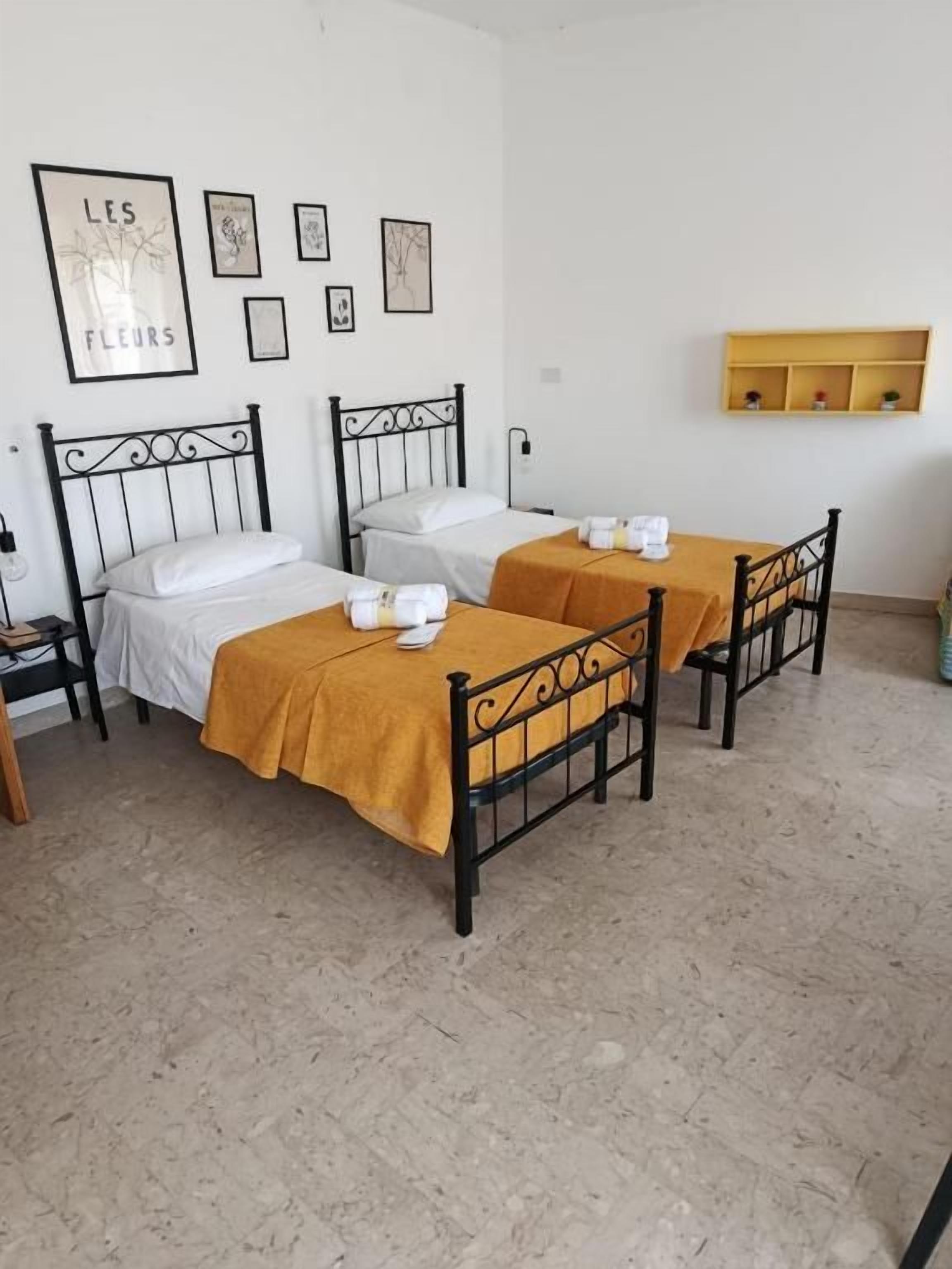 Une chambre avec deux lits, chacun doté d’une tête de lit et d’une table de chevet. Des tableaux encadrés ornent le mur, et une étagère se trouve au-dessus des lits.