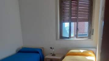 A room with two beds, one with a blue bedspread and the other with an orange bedspread. There is a window with blinds, a small table with a lamp, and a wall-mounted light fixture.