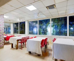 A dining area with tables covered in white tablecloths and red chairs, large windows with a view of greenery outside. - Deluxe Aysha Stay Inn (Kodaikanal)