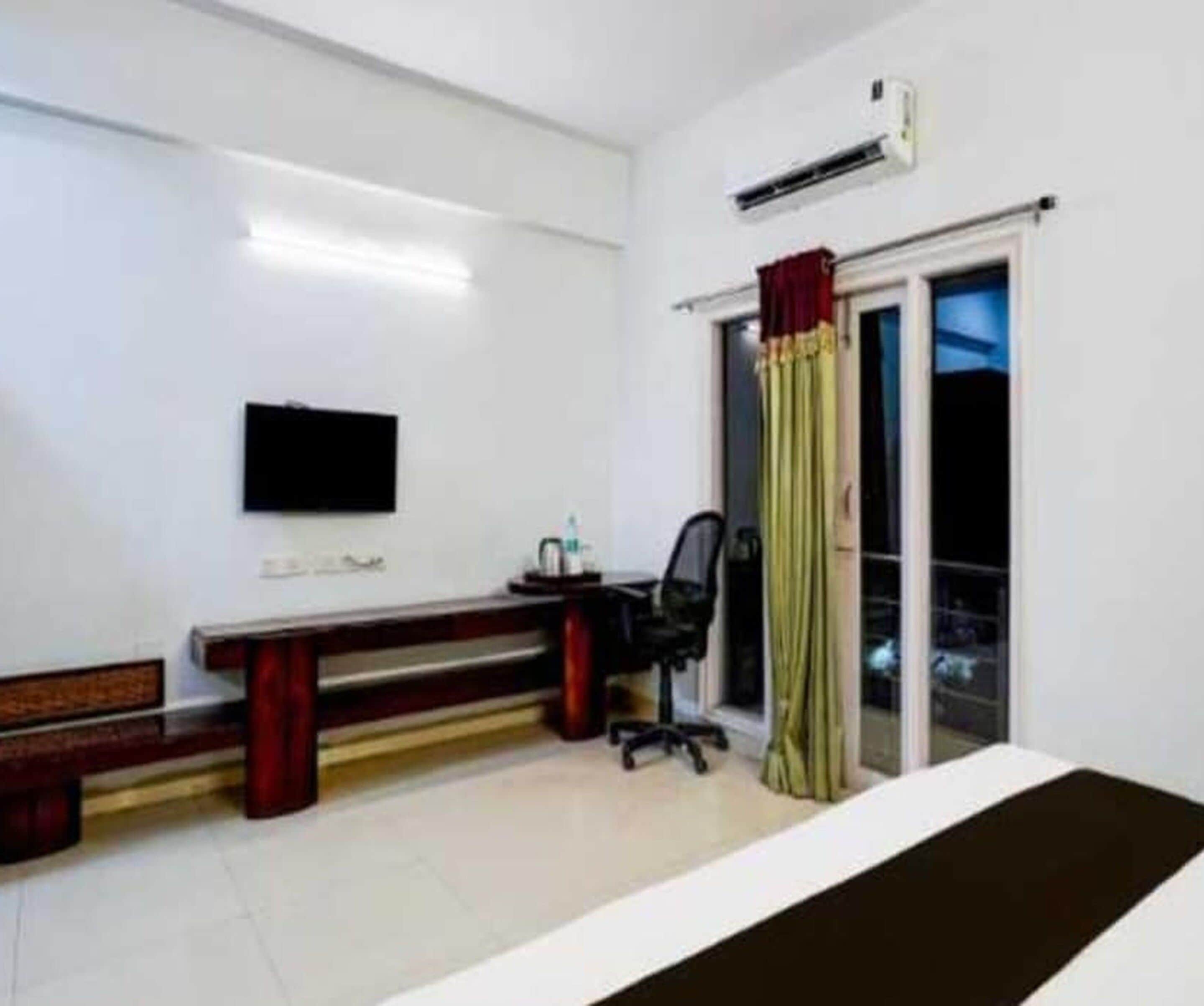 A hotel room with a bed, a desk with a chair, a flat-screen TV, and a window with curtains.