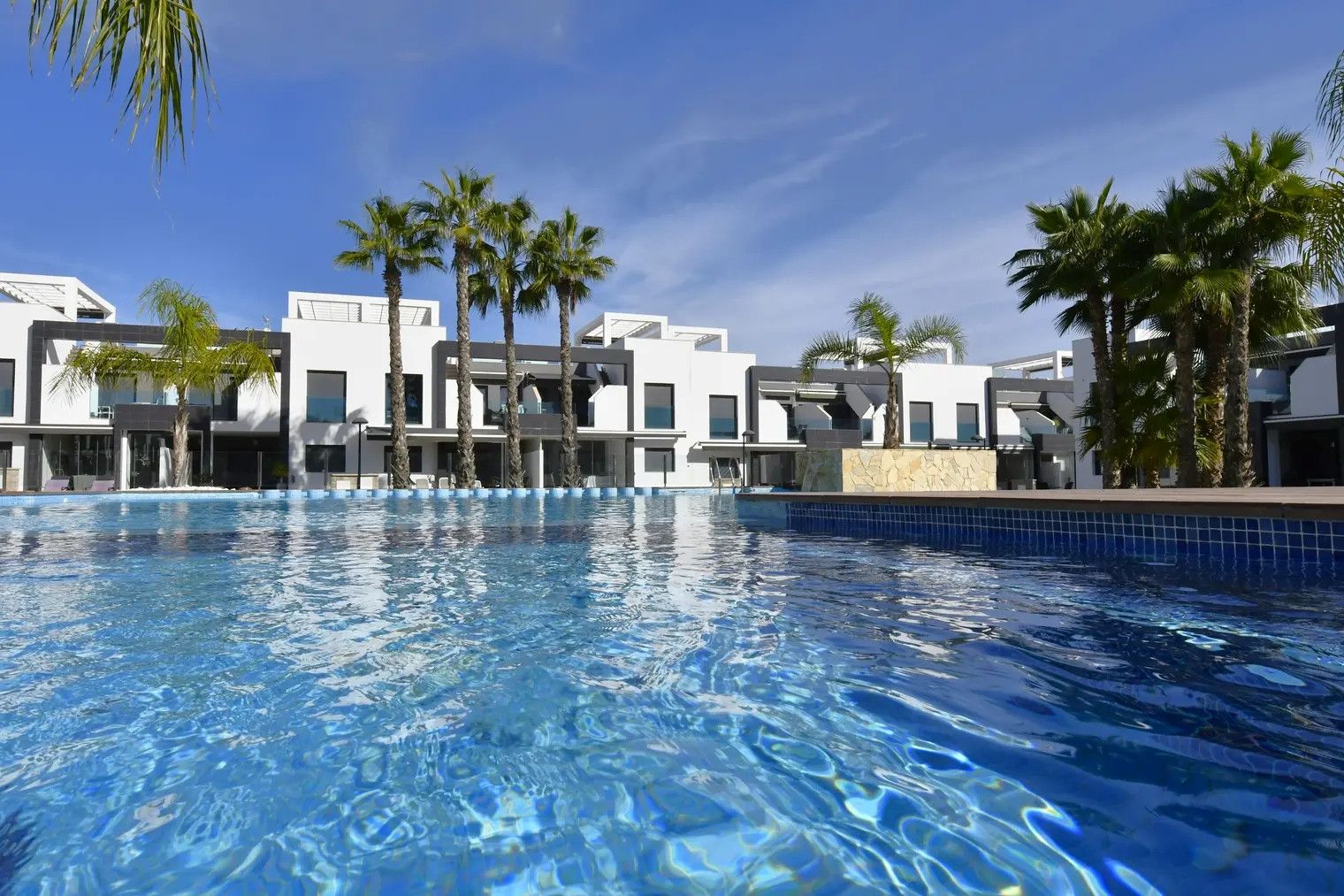A swimming pool with clear blue water, surrounded by modern buildings with white facades and palm trees.