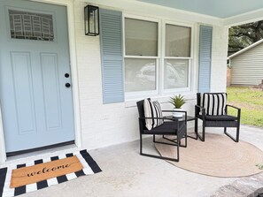 Un porche à l’avant de la maison, avec une porte bleue, un paillasson, deux chaises, une petite table et un pot de fleurs.