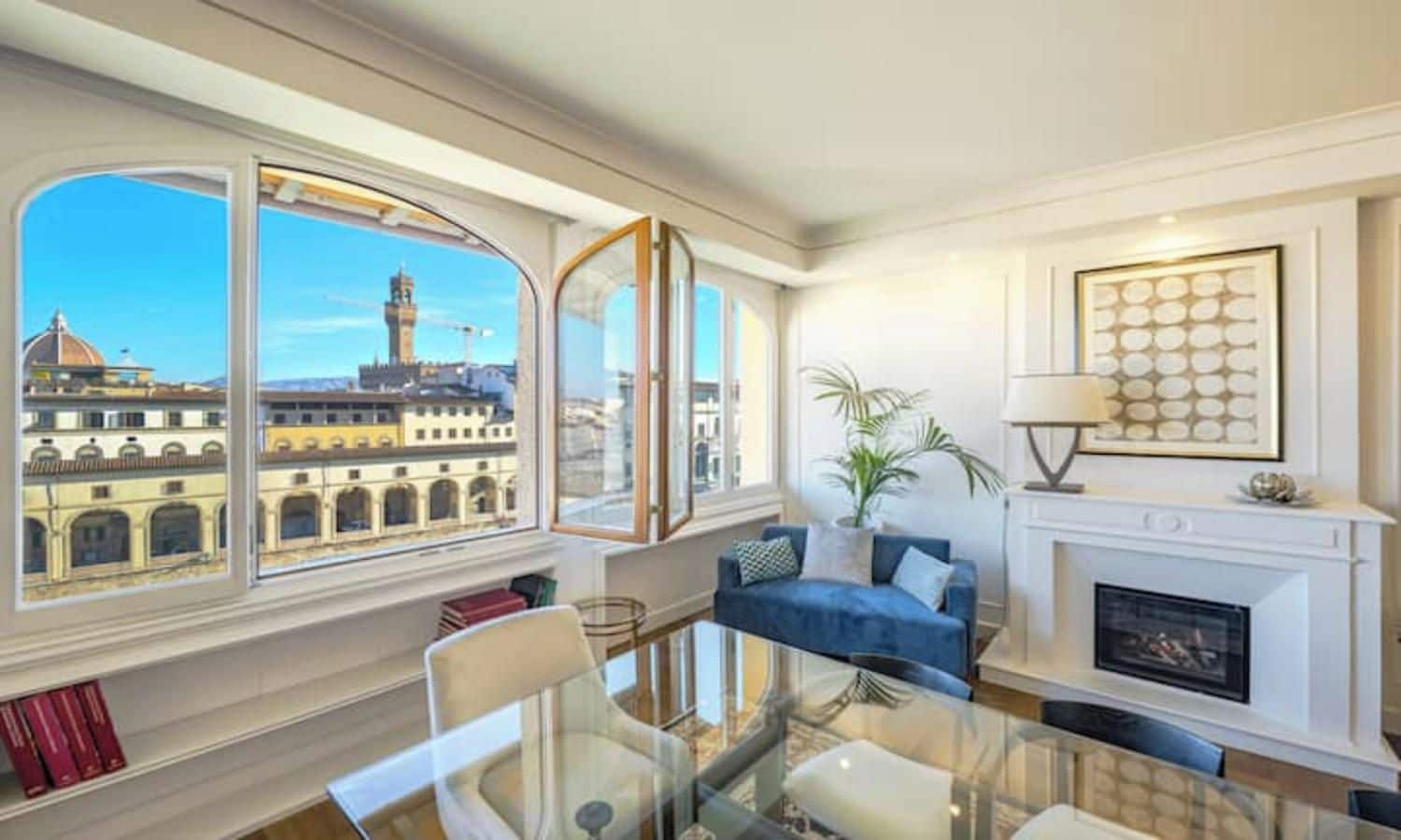 A room with a glass table, a blue sofa, a fireplace, and a view of a cityscape with historical buildings.