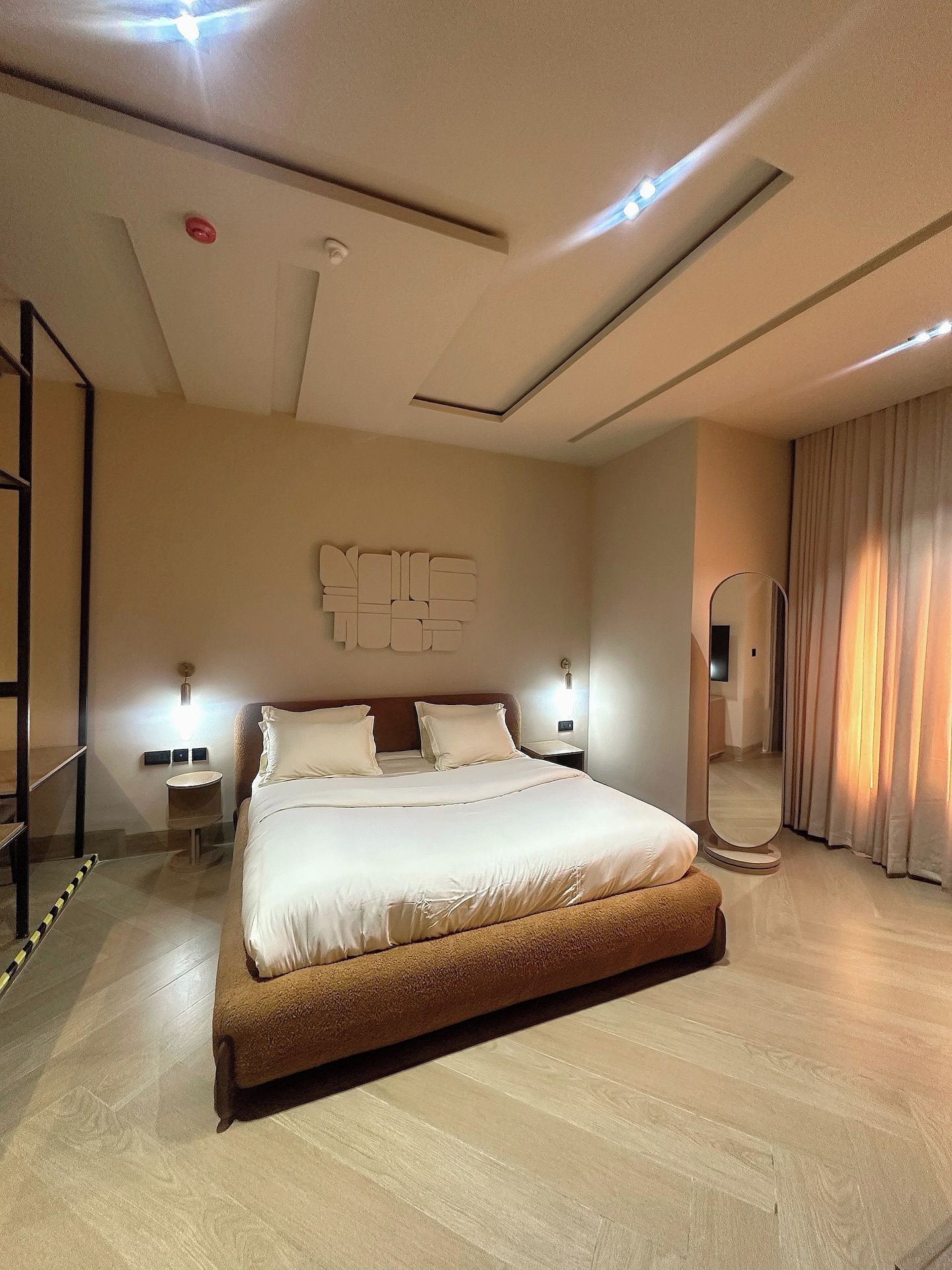 A modern hotel room with a large bed, a mirror, a wall-mounted light fixture, and a ceiling light.