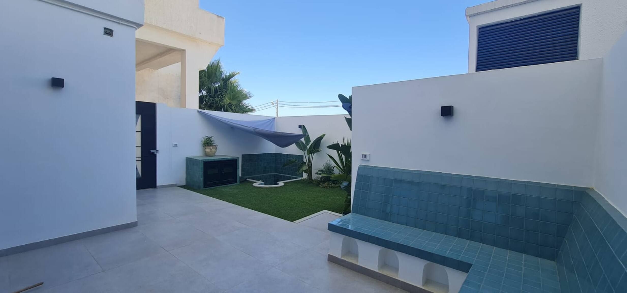 Un espace patio extérieur moderne, doté d’un banc carrelé, d’un petit jardin avec gazon synthétique et d’un mur recouvert de carreaux bleus.