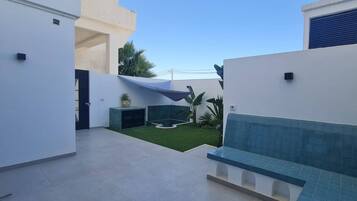 Un espace patio extérieur moderne, doté d’un banc carrelé, d’un petit jardin avec gazon synthétique et d’un mur recouvert de carreaux bleus.