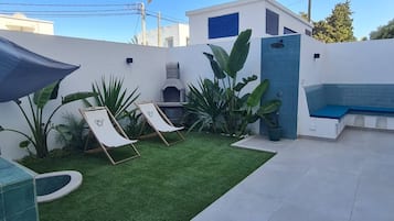 Uma área de pátio com grama sintética, duas poltronas de descanso, uma pequena piscina e um banco.