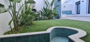 Un patio con césped artificial, una piscina con azulejos y diversas plantas.