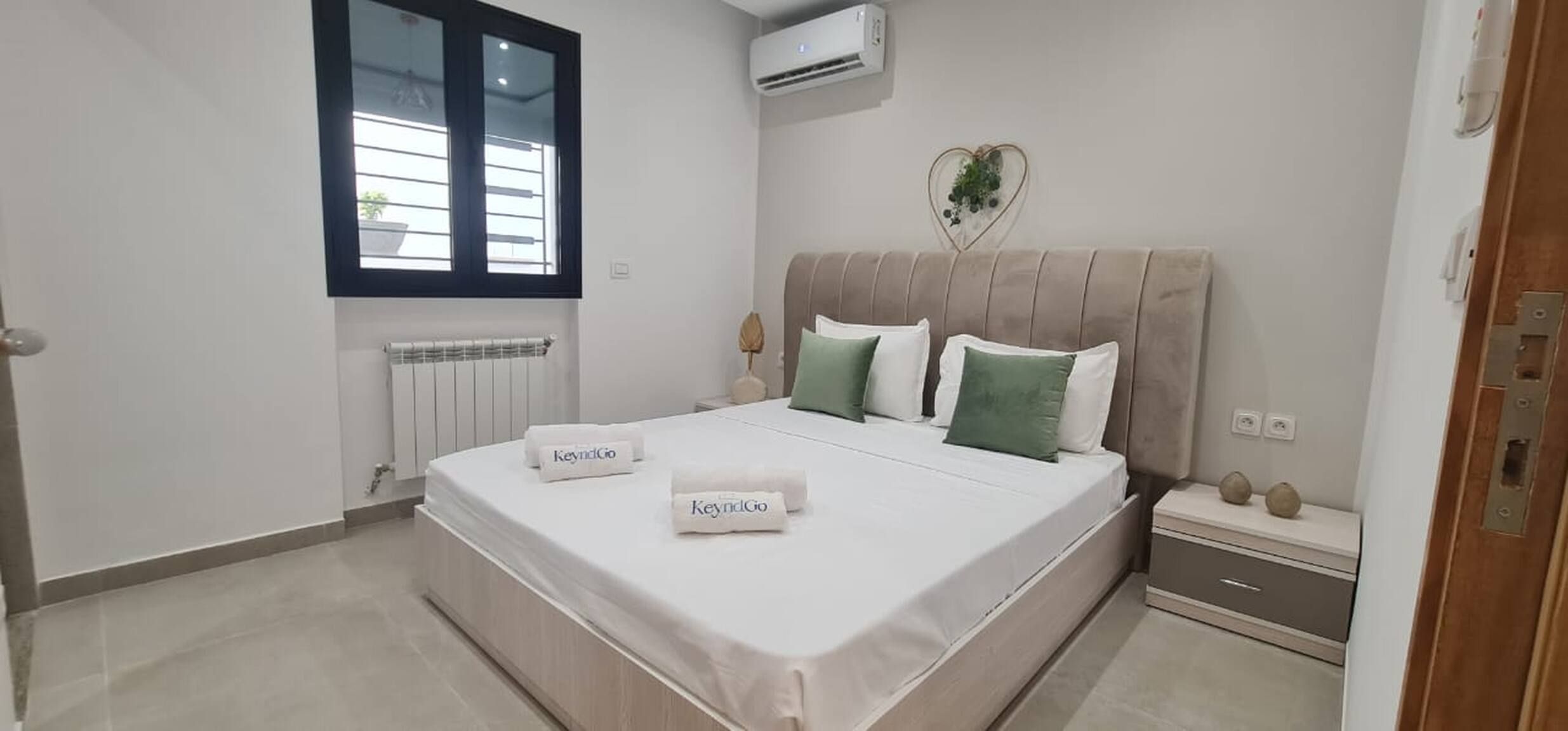 Ein Schlafzimmer mit Bett, Fenster, Klimaanlage, Heizkörper, Nachttisch und Beistelltisch.