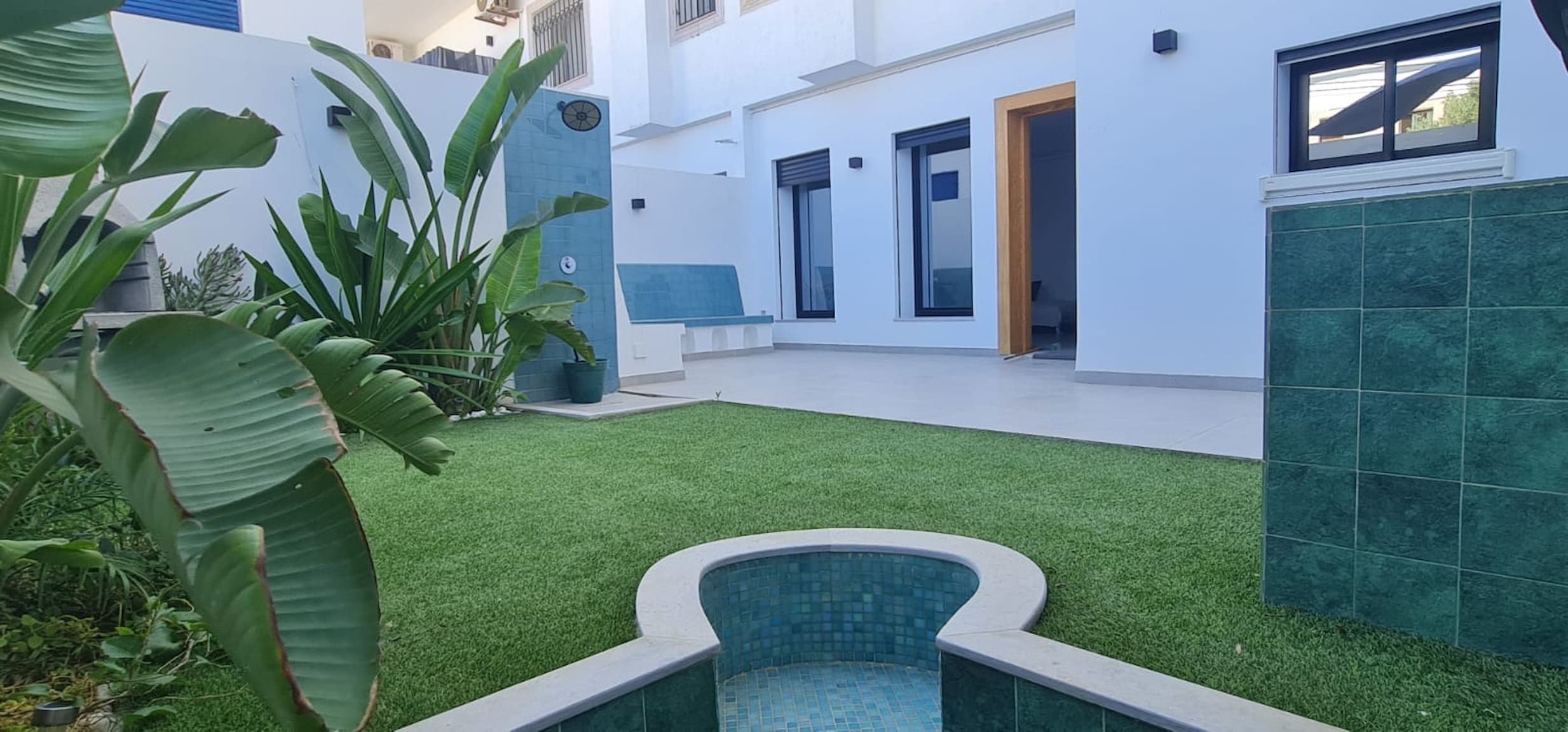 Un edificio moderno con piscina, césped artificial verde y plantas tropicales.