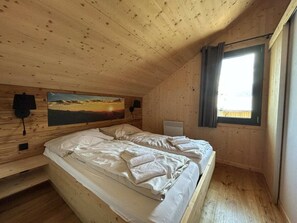 A wooden cabin bedroom with a bed, a painting, a heater, and a window with curtains. - One-bedroom Cabin (Sankt Georgen am Kreischberg)