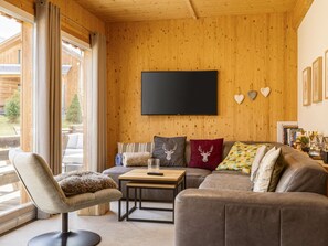 Ein gemütliches Wohnzimmer mit Holzwand, einem Flachbildfernseher, einer Couch, einem Sessel und einem Couchtisch.