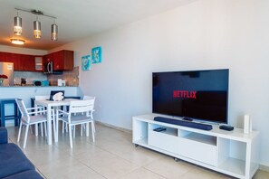 Ein modernes Wohnzimmer mit einem Flachbildfernseher, der 'NETFLIX' zeigt, einem Essbereich mit weißen Stühlen, einer Küche mit roten Schränken und einer blauen Akzentwand.