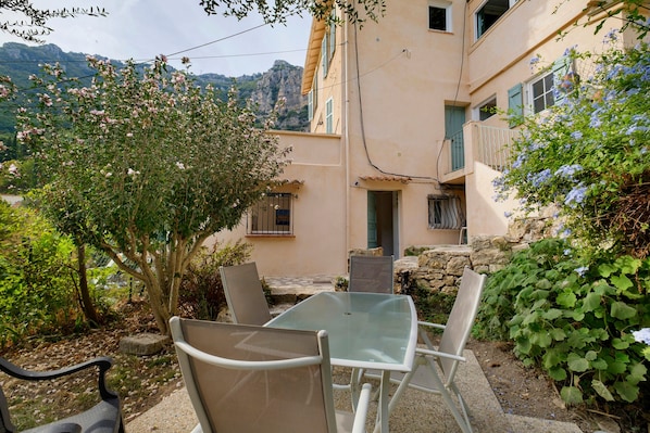 Un pati amb taula i cadires de vidre, envoltat de verdura i un edifici amb mur de pedra.