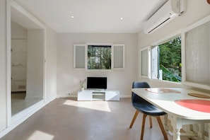Habitació moderna amb un televisor de pantalla plana sobre un suport blanc, una taula de menjador amb caires i una finestra amb vista a vegetació.