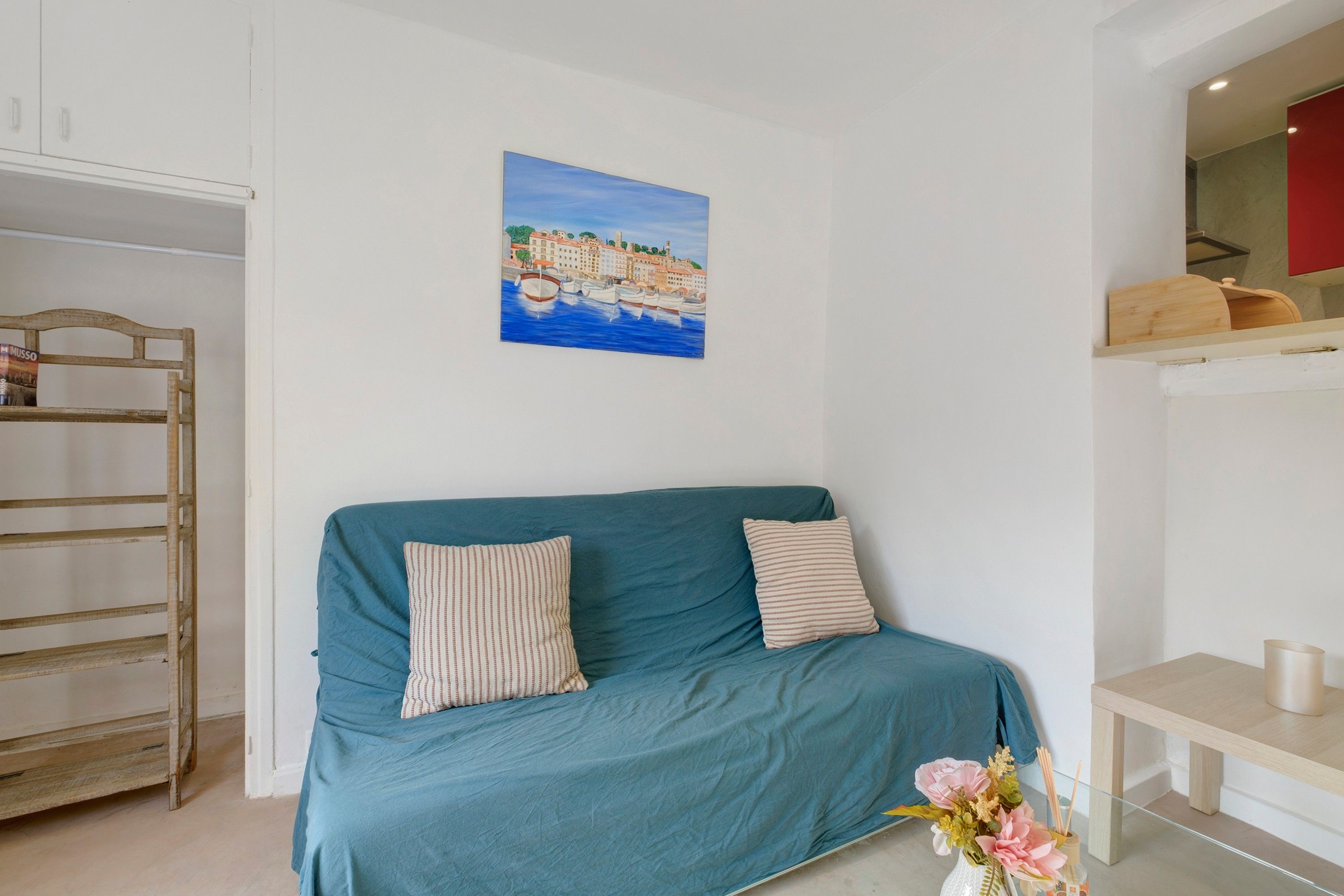 Ein Zimmer mit einem petrolfarbenen Sofa, einem Holzstuhl, einem Gemälde mit einer Küstenlandschaft und einem kleinen Tisch mit einem Blumenstrauß.