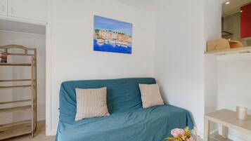 Ein Zimmer mit einem petrolfarbenen Sofa, einem Holzstuhl, einem Gemälde mit einer Küstenlandschaft und einem kleinen Tisch mit einem Blumenstrauß.