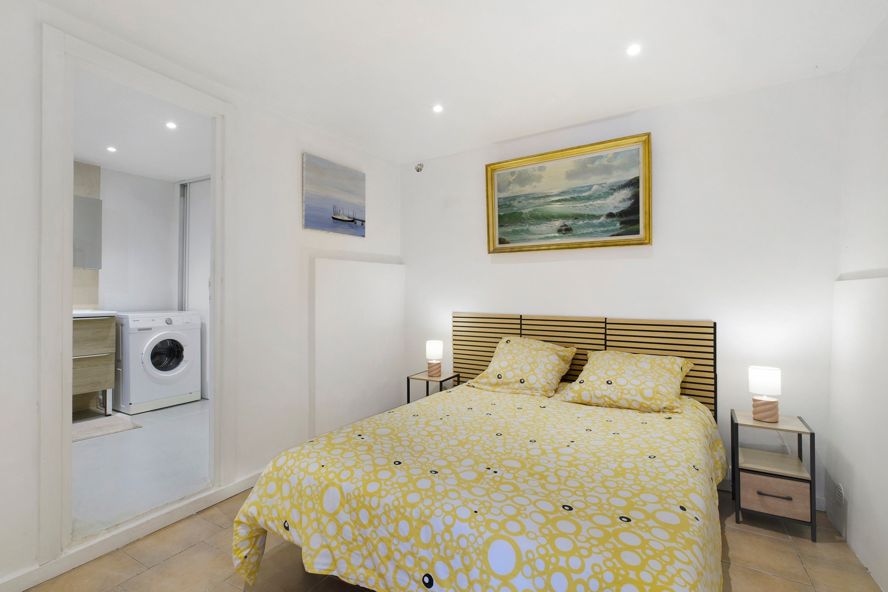 Een slaapkamer met een bed, een wasmachine, een schilderij en een nachtkastje.