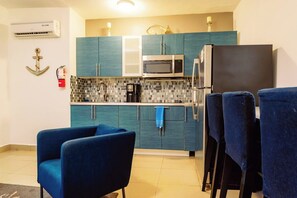 Eine kompakte Küche mit türkisgrünen Schränken, einer Mikrowelle und einem Kühlschrank. Im Vordergrund stehen zwei blaue Stühle.