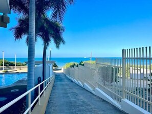 A pathway leading to a pool with a view of the ocean, surrounded by a white fence and palm trees. - Beachside Condo with Pool and Ocean Views (Pole Ojea)