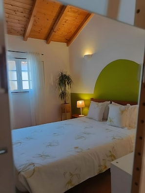 Een slaapkamer met een bed, nachtkastje, lamp, een pot met een plant en een raam met gordijnen.