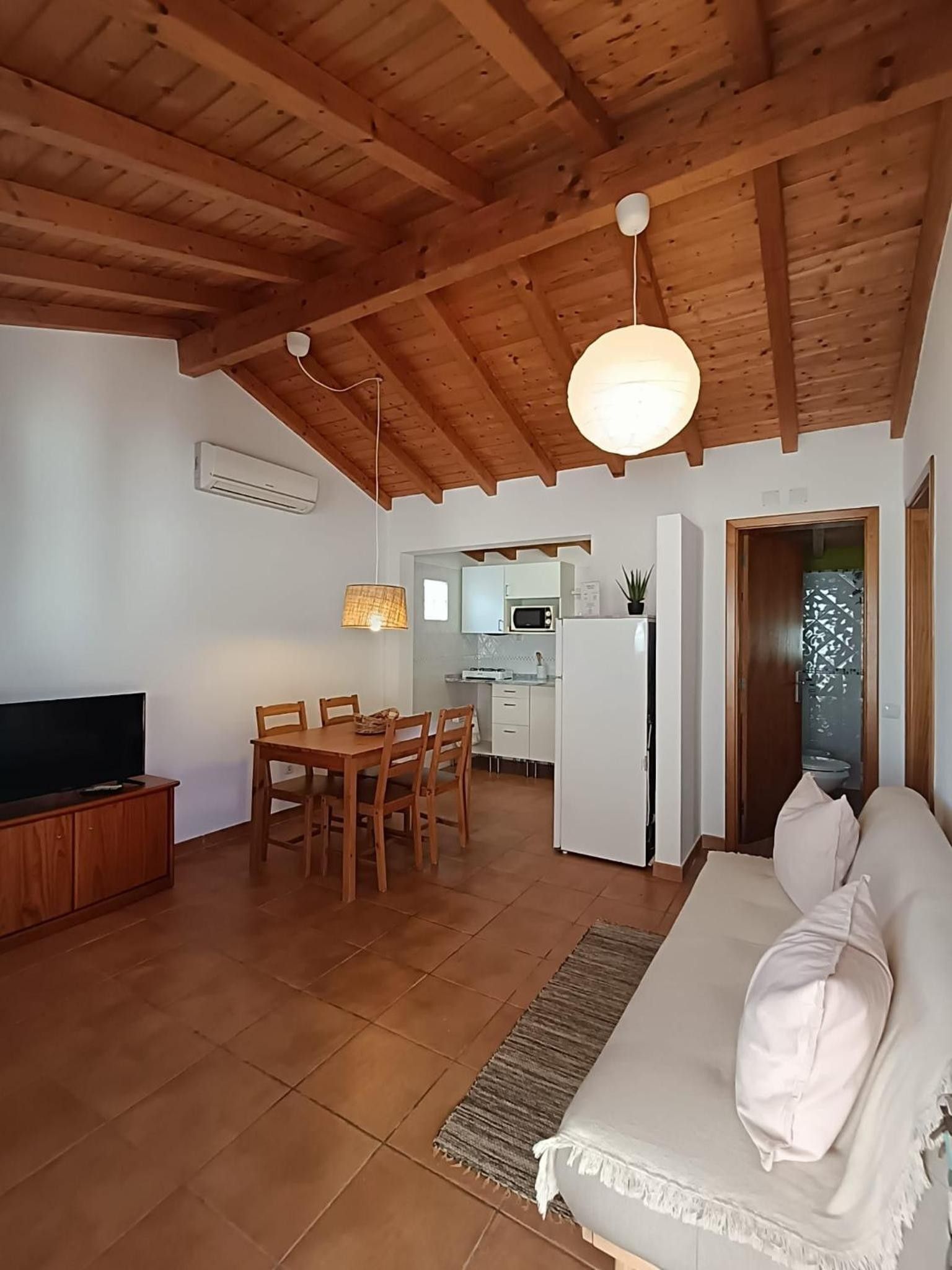 Un salon avec un plafond en bois, un coin repas avec une table et des chaises, une cuisine avec des meubles blancs et un canapé agrémenté de coussins.