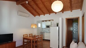 Ein Wohnzimmer mit Holzbalkendecke, eine Essecke mit Tisch und Stühlen, eine Küche mit weißen Schränken sowie ein Sofa mit Kissen.