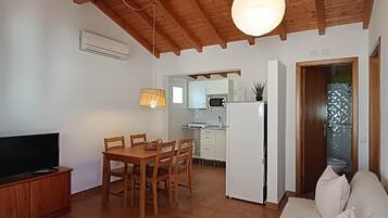 Un salon avec un plafond en bois, un coin repas avec une table et des chaises, une cuisine avec des meubles blancs et un canapé agrémenté de coussins.