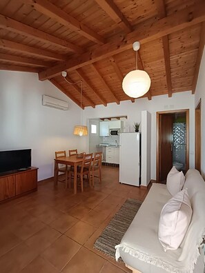 Uma sala de estar com teto de madeira, uma área de jantar com mesa e cadeiras, uma cozinha com armários brancos e um sofá com almofadas.