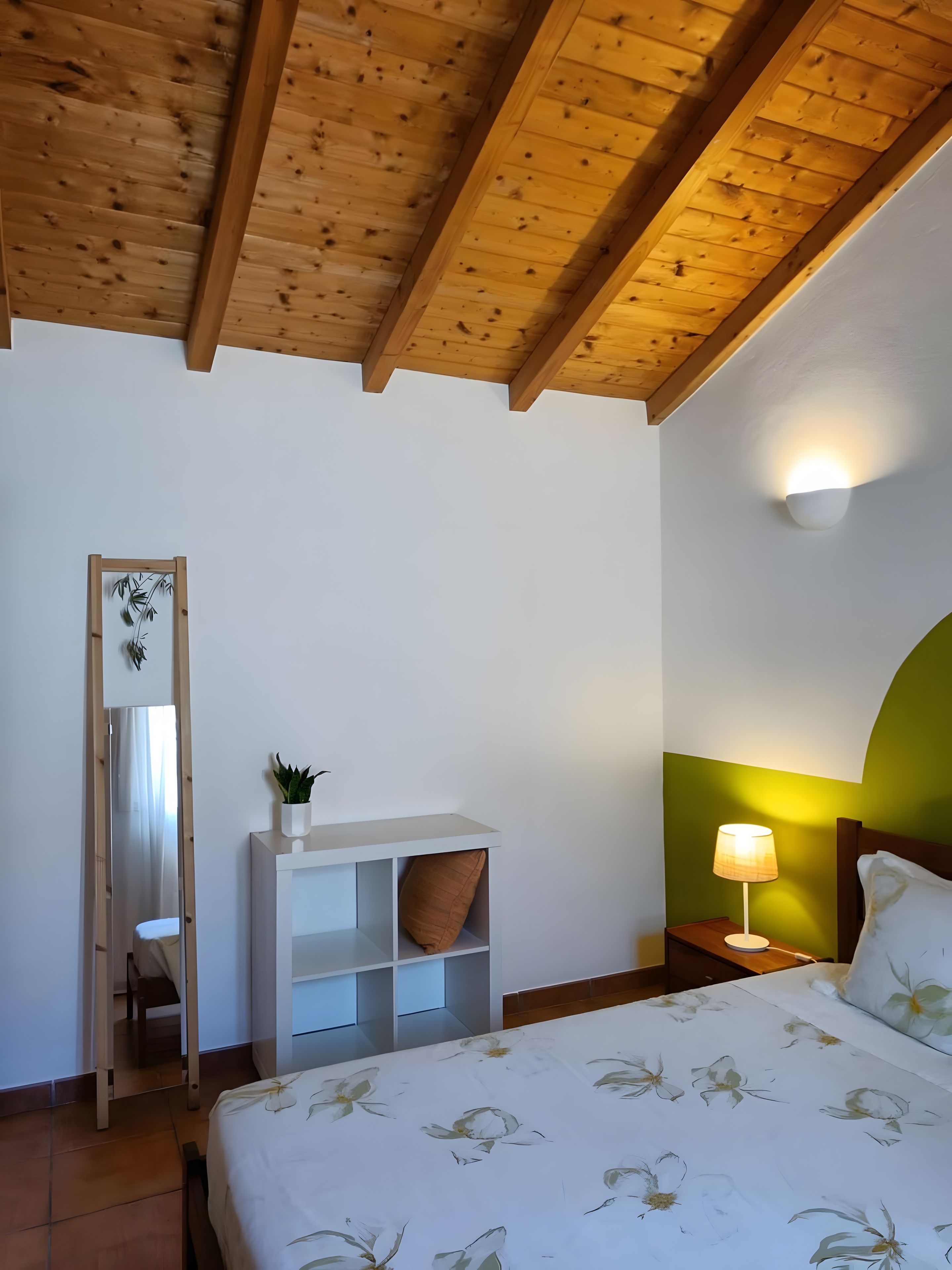 A bedroom with a wooden ceiling, a bed with floral bedding, a nightstand with a lamp, and a mirror with a plant.