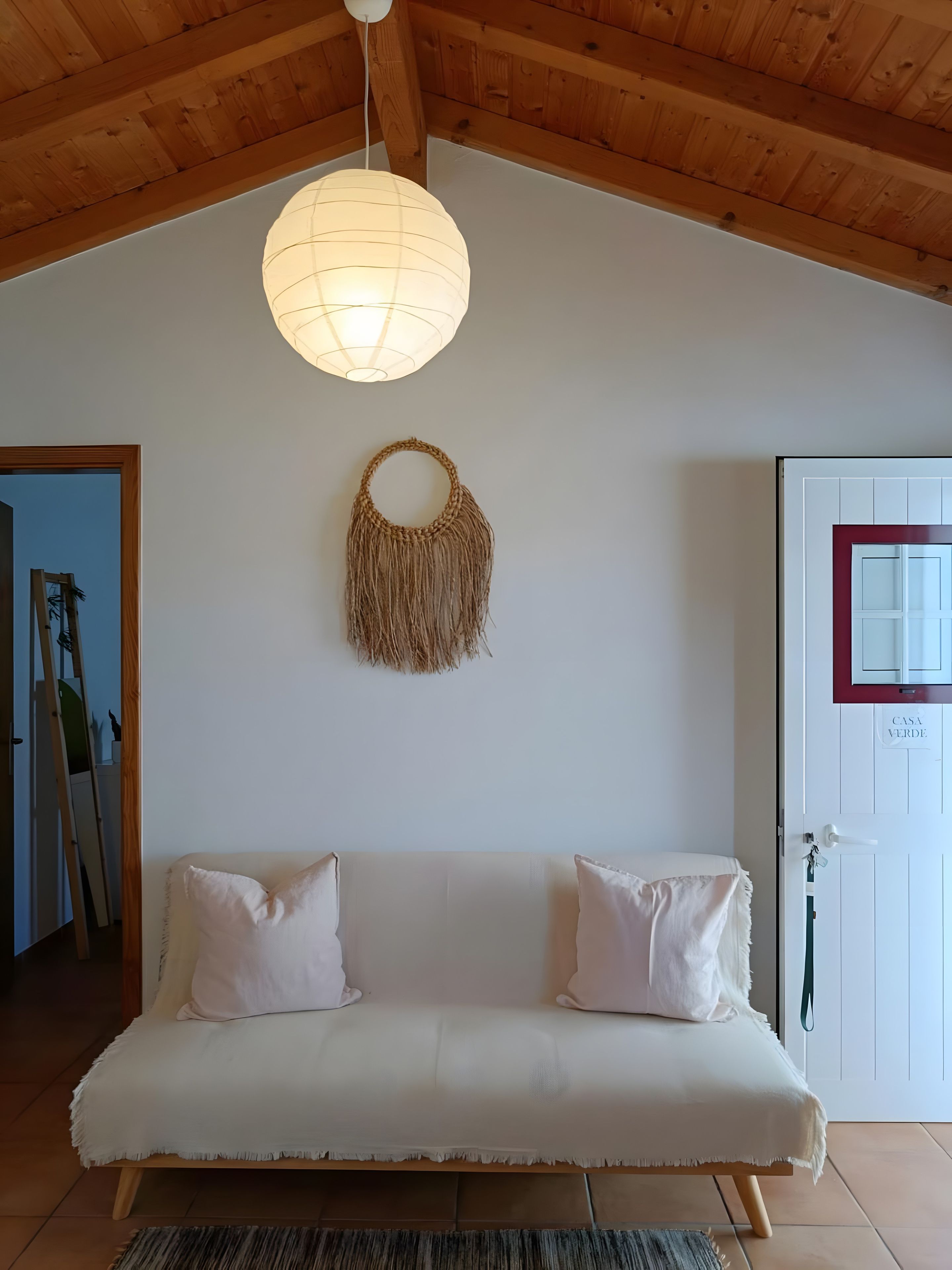 Une pièce chaleureuse avec un plafond en bois, une décoration murale en macramé suspendue, un canapé blanc agrémenté de deux coussins et une porte avec une pancarte.