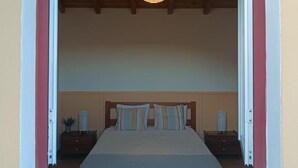 A bedroom with a bed, bedside tables, a hanging light fixture, and a window with a wooden frame. - Apartment 'Casa Medronheiro' with Mountain View, Wi-Fi and Air Conditioning (Melides)