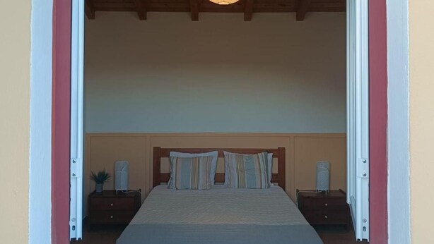 Un dormitori amb un llit, taules de nit, una làmpada penjant i una finestra amb marc de fusta.