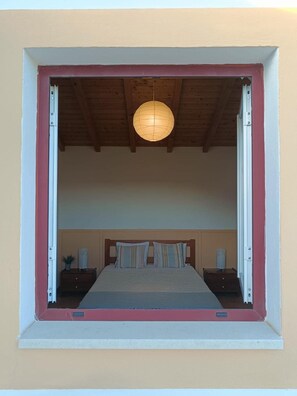 Quarto com cama, mesas de cabeceira, luminária pendente e janela com moldura de madeira.