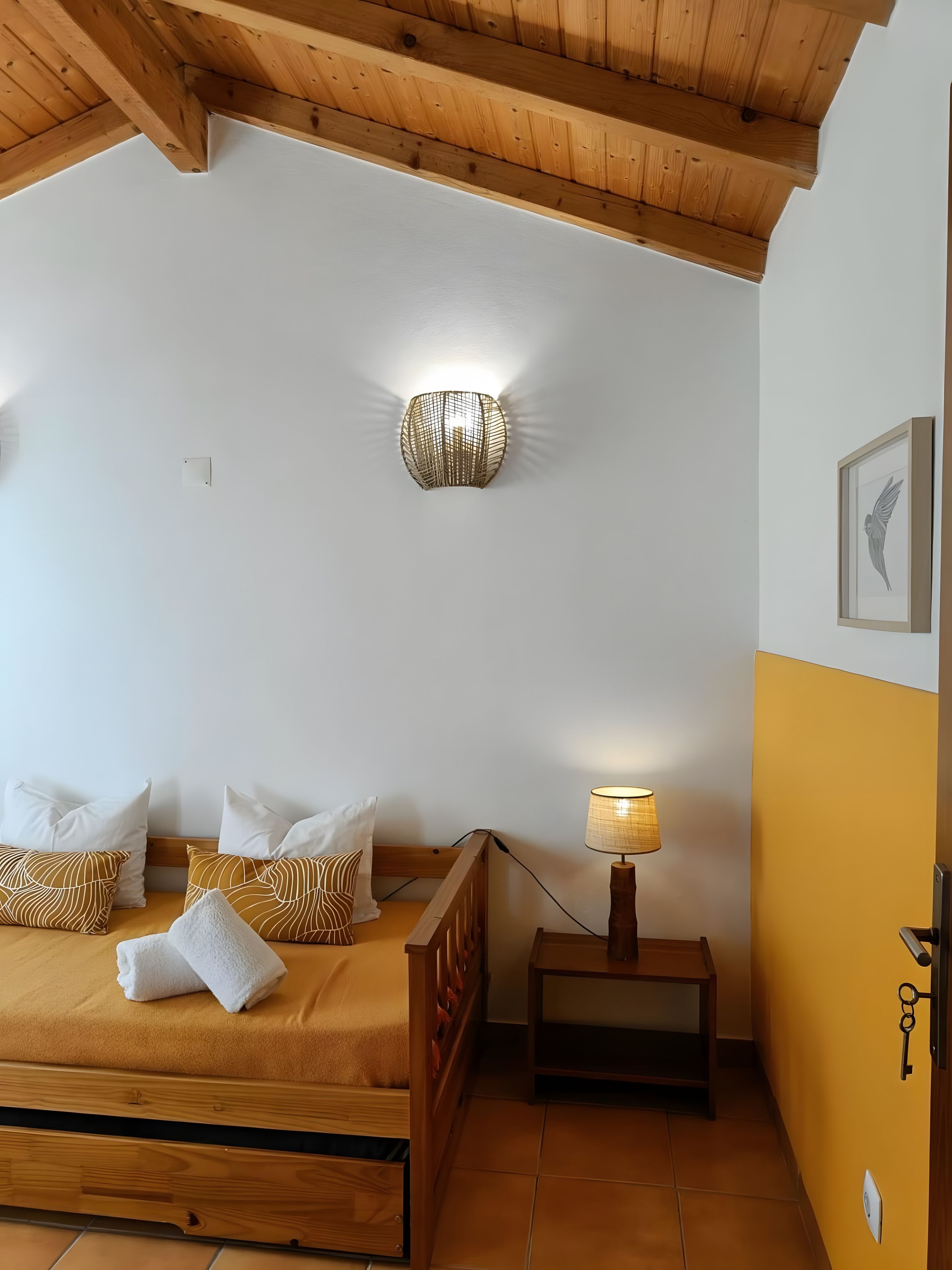 A bedroom with a wooden bed, white and patterned pillows, a wall-mounted light fixture, a bedside table with a lamp, and a framed picture on the wall.