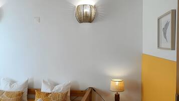 A bedroom with a wooden bed, white and patterned pillows, a wall-mounted light fixture, a bedside table with a lamp, and a framed picture on the wall.