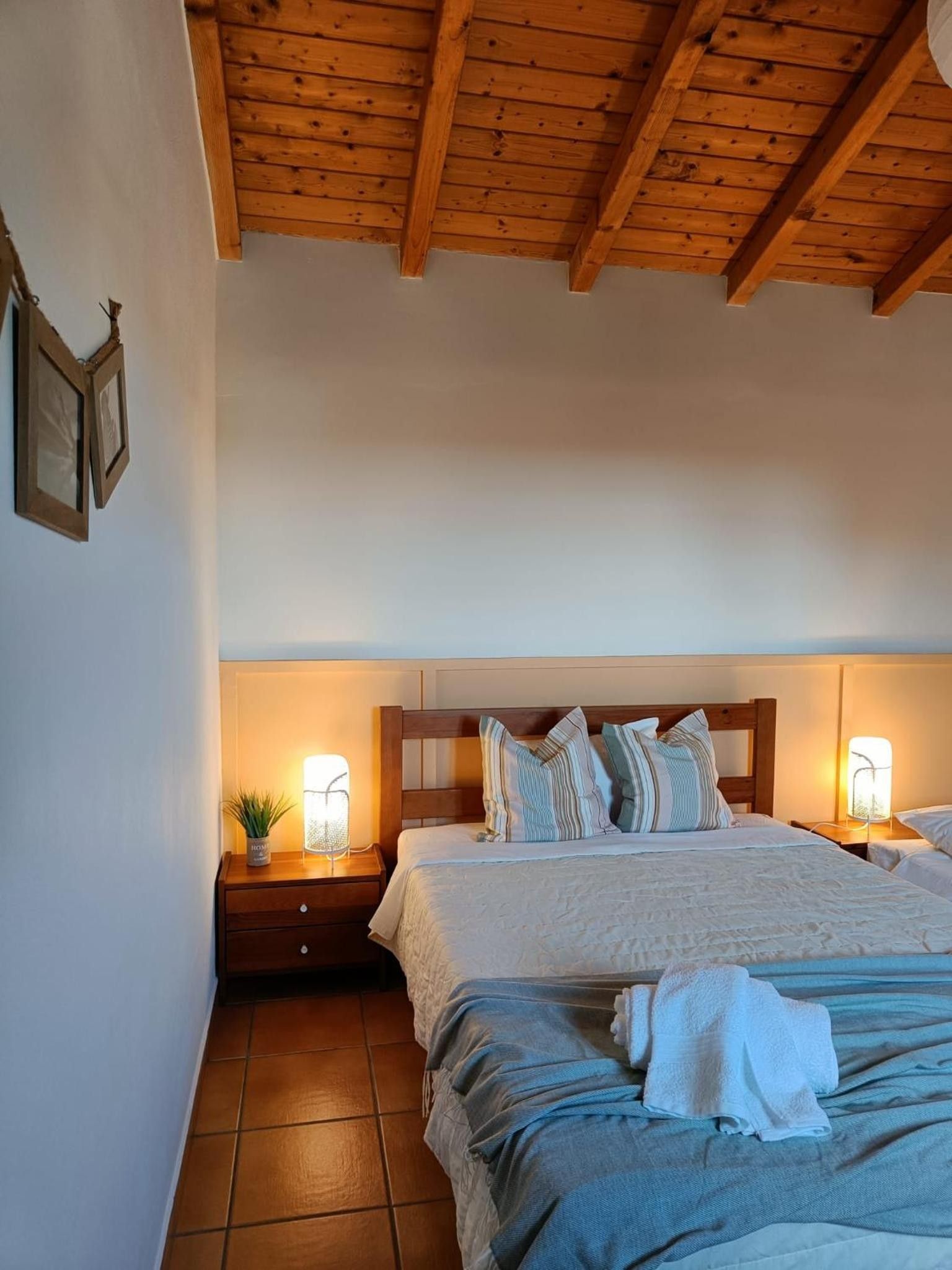 A bedroom with a wooden ceiling, a bed with pillows, a nightstand with a lamp, and a wall-mounted picture.