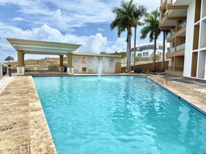 A swimming pool with a fountain, surrounded by a building with balconies and palm trees. - Family-Friendly | Year-Round Pool & Beach Nearby (Boquerón)