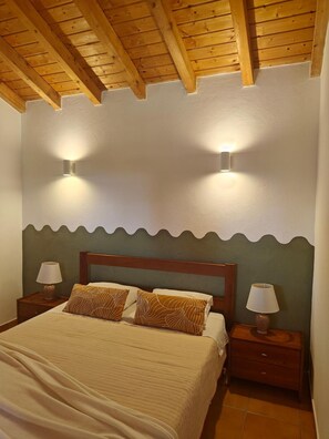 Un dormitori amb sostre de fusta, dues làmpades penjades a la paret, un llit amb coixins estampats i tauletes de nit amb llums.