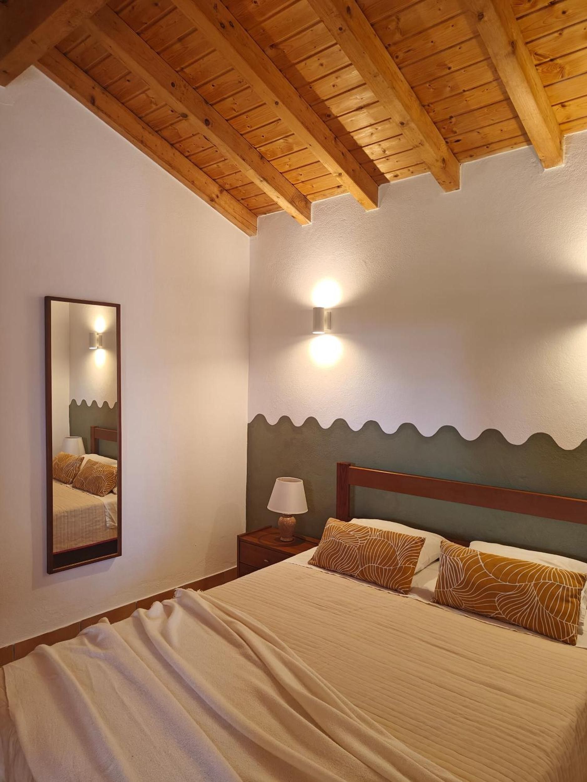 Une chambre avec un plafond en bois, un lit agrémenté d’oreillers à motifs, une table de chevet avec une lampe et un luminaire fixé au mur.