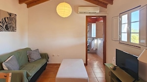 A cozy living room with a wooden ceiling, a green sofa, a white ottoman, a television, and a hanging lamp. - Apartment 'Casa Laranjeira' with Mountain View, Wi-Fi and Air Conditioning (Melides)