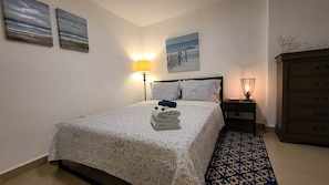 Um quarto com cama, mesa de cabeceira, luminária, cômoda e quadros nas paredes.