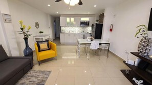 Un salon moderne avec un canapé, un fauteuil jaune, une table à manger et une cuisine en arrière-plan.