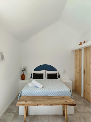 Quarto com cama, mesas de cabeceira, um banco de madeira, um vaso com planta e guarda-roupa.